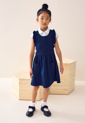 Ragazza giovane con chignon, che indossa un vestito blu navy con volant, blusa bianca, calzini svolazzanti e scarpe nere, in piedi accanto a blocchi di legno.