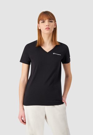 Jonge vrouw met rood haar, gekleed in een zwart Champion V-hals T-shirt en lichtgekleurde broek, tegen een effen grijze achtergrond.