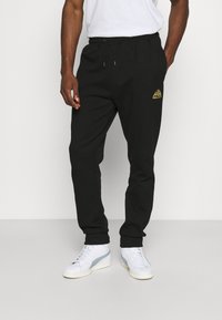 Black joggers with an elastic waistband and drawstring, featuring a gold logo on the left thigh. Styled with white sneakers.