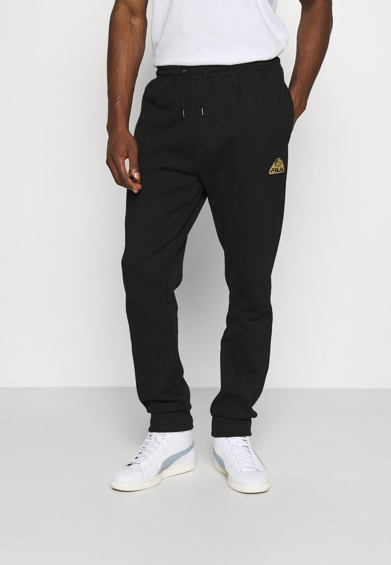 Black joggers with an elastic waistband and drawstring, featuring a gold logo on the left thigh. Styled with white sneakers.