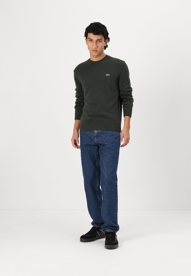 Dunkelgrüner Pullover mit gerippten Bündchen und Saum, mit einem kleinen Logo. Kombiniert mit blauen Jeans und schwarzen Sneakern.