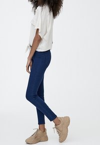 Jean skinny en denim bleu foncé, associé à un haut léger à manches courtes noué à la taille, et des baskets beiges à semelles texturées.