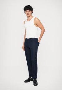 Calças azul-marinho em corte reto com uma textura suave. Combinadas com uma t-shirt branca e sapatos pretos, mãos nos bolsos.