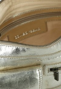 Metallisch silbernes Handtascheninnere mit einer glatten Textur, ausgestattet mit Reißverschlussfächern und einem beigen Futter mit geprägtem Branding.