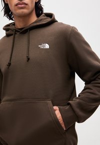 Homme portant un sweat à capuche marron avec une poche avant et le logo The North Face sur la poitrine gauche, main partiellement dans la poche.