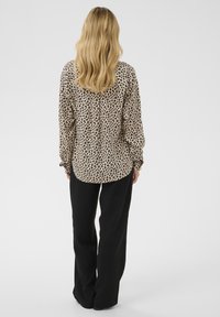 Pikade blondidega naine seisab seljaga, kandes beežikat leopardimustriga pluusi ja lahtisi musti pükse, taust on ühtlane.