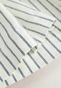 Tissu blanc avec des fines rayures verticales bleu marine régulièrement espacées et des coutures visibles, plié sur une surface plane.