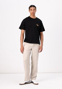 T-shirt noir avec un logo doré sur la poitrine, associé à un pantalon beige clair et des baskets blanches avec des accents noirs.