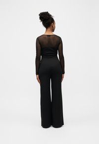 Femme aux cheveux bouclés en chignon portant un haut noir transparent à manches longues et un pantalon noir taille haute à jambes larges, debout, de dos.