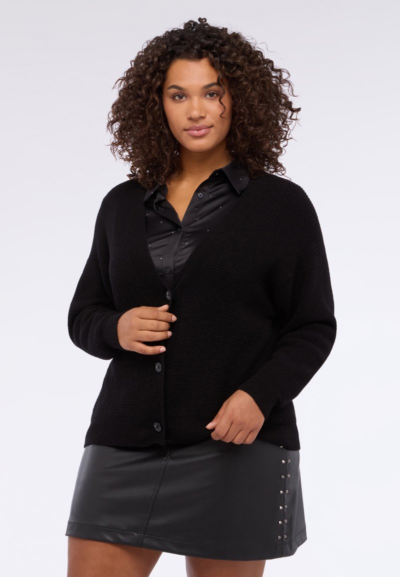 Cardigan nero lavorato a maglia con scollo a V e chiusura con bottoni, indossato sopra una camicetta di raso nera. L'outfit è abbinato a una gonna in pelle nera.