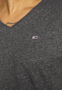 Ciemnoszara koszulka V-neck z miękkiego, teksturowanego materiału, z małym haftowanym logo w kolorach czerwonym, białym i niebieskim na piersi.