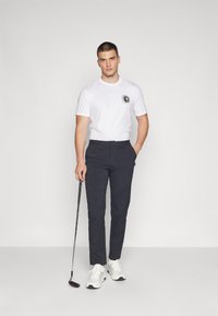 Witte kortlopende t-shirt met grafische print, marineblauwe pantalon, en witte sportschoenen. Man houdt een golfclub vast.
