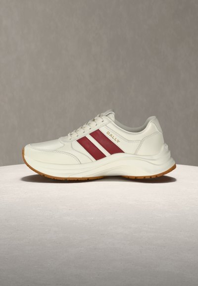 Baskets basses blanches avec des rayures latérales rouges, le logo "BALLY" doré et une semelle en gomme, présentées sur une surface claire avec un fond gris.