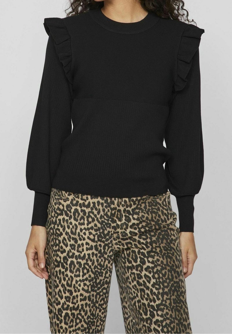 Femme portant un pull noir à manches longues avec des volants aux épaules et un pantalon imprimé léopard, debout devant un fond gris.