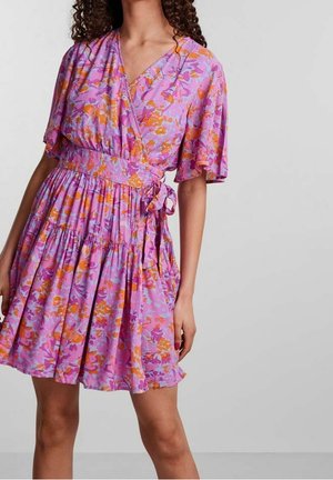 Femme portant une robe portefeuille florale rose et orange avec de courtes manches volantées et un nœud à la taille, se tenant devant un fond gris uni.