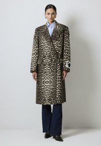 Femme portant un long manteau en imprimé léopard, une chemise bleu boutonnière, un jean évasé noir et des chaussures noires à bouts pointus sur un fond clair uni.