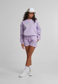 Lila bomullströja med "Kani"-logotyp, matchande shorts, vita sneakers och en vit keps med nätbak. Slät textur, avslappnad design.