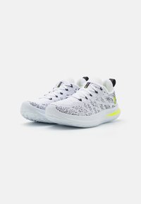 Une paire de chaussures de sport blanches et noires tachetées avec des semelles blanches et des accents jaunes fluo, présentée sur un fond blanc.