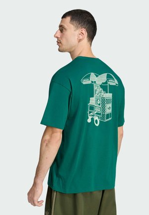 Uomo con t-shirt verde Adidas con disegno a linee bianche di un carretto di venditore ambulante e scarpe da ginnastica sulla schiena, in piedi davanti a uno sfondo semplice.