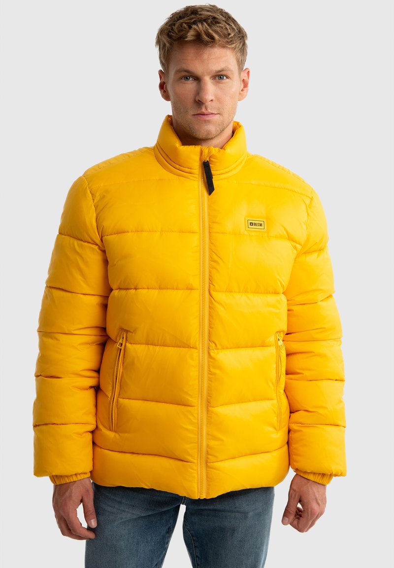 Geltona pūkuota striukė su aukštu apykakle, su horizontaliais siuvinėti raštais, užtrauktukais kišenėse ir logotipu ant krūtinės.