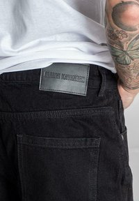 Los jeans de denim negro presentan un parche de cuero con "GIANNI KAVANAGH" grabado. La tela tiene una textura suave y costuras distintivas.