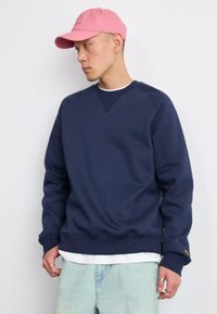 Marineblå sweatshirt med rund hals og ribbede manchetter, lagd over en hvid skjorte. Lyseblå denimbukser. Lyserød kasket med sort tekst.