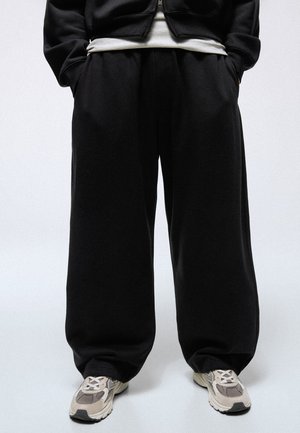 Persona che indossa pantaloni neri larghi, scarpe da ginnastica bianche e grigie, con le mani in tasca su uno sfondo chiaro uniforme.