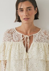 Spitzenbluse in Creme mit Volantdesign, Rundhalsausschnitt und Schleifenverschluss vorne. Filigranes Blumenmuster überall.