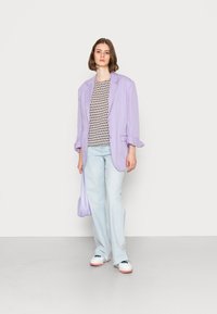 Blazer oversized lavanda sobre uma blusa estampada, combinado com jeans claros de boca de sino e ténis brancos com detalhes em rosa.