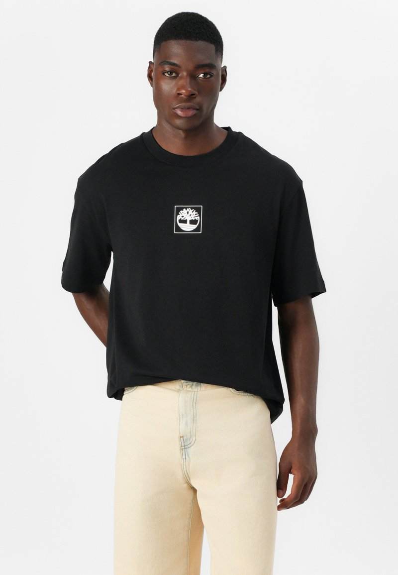 Czarna koszulka z krótkim rękawem z białym logo drzewa na środku. Materiał jest gładki, luźny krój. Połączona z beżowymi spodniami o prostym kroju.