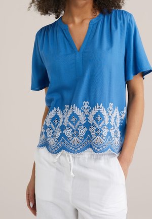 Femme portant un chemisier bleu à manches courtes avec une broderie florale blanche à l'ourlet et un pantalon blanc avec cordon de serrage.