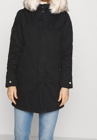 Svart parka med huva i fuskpäls, tryckknappar och dragkedja, sidofickor och ribbade cuffs. Har en något överdimensionerad passform.