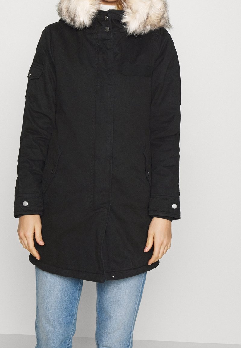 Svart parka med huva i fuskpäls, tryckknappar och dragkedja, sidofickor och ribbade cuffs. Har en något överdimensionerad passform.