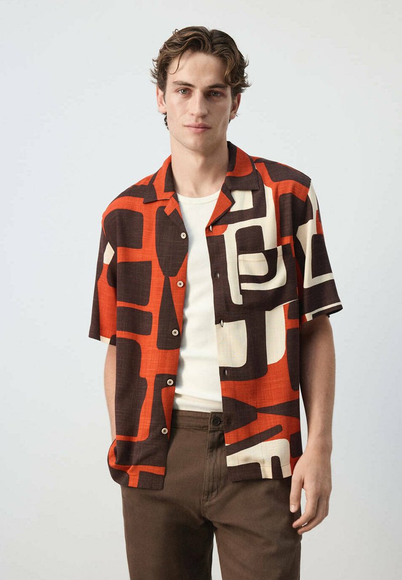 Jeune homme portant une chemise à manches courtes à boutons avec un motif abstrait orange, marron et crème, associée à un pantalon marron et un sous-vêtement blanc.