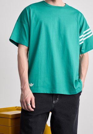 Personne portant une chemise à manches courtes couleur sarcelle avec trois bandes blanches sur la manche gauche et un petit logo près de l'ourlet, associée à un pantalon noir.