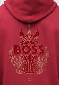 Felpa con cappuccio rossa con un evidente logo "BOSS", ricamo dorato dettagliato e un elemento a trama vicino allo scollo.