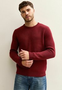 Czerwony sweter o strukturze z okrągłym dekoltem, ściągaczami na rękawach i u dołu, noszony ze niebieskimi dżinsami. Wzór dzianiny dodaje głębi i zainteresowania.