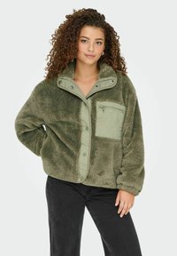 Vrouw met krullend haar, gekleed in een olijfgroene fleecejas met ritssluiting op de borstzak, gecombineerd met een zwarte broek, staand tegen een lichte achtergrond.