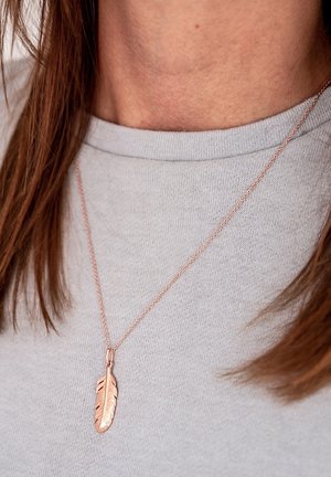 Rosen goldene Halskette mit einem federförmigen Anhänger, der mit strukturierten Details versehen ist, getragen über einem hellgrauen Shirt mit Rundhalsausschnitt.