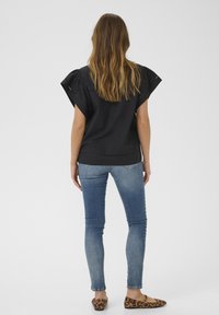 Fekete, rövid ujjú felső, gyűrt vállakkal, szűk kék farmernadrággal és leopárdmintás balerinacipővel párosítva, hátulról nézve.