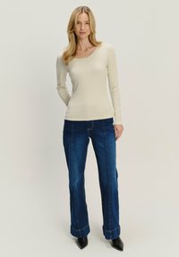 Maglietta a maniche lunghe color crema a costine abbinata a jeans a zampa d'elefante blu scuro con dettagli di cucitura e décolleté neri a punta. Sfondo chiaro.