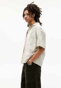 Ung person med dreadlocks iført en kortærmet, ternet skjorte med knapper og mørke bukser, som står med den ene hånd i lommen.