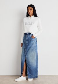 Ženska v beli srajci z dolgimi rokavi z napisom "BOSS", modrih visokih jeans krilu maxi z odprtinami spredaj in belih teniskah, stoji.