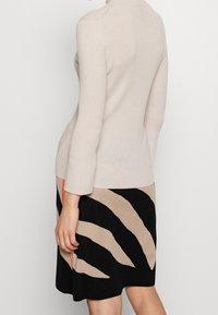 Donna che indossa un top beige a maniche lunghe e una gonna nera con ampie strisce astratte beige, in piedi su uno sfondo bianco semplice.