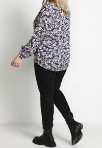 Frau, die eine blumige Langarmbluse, schwarze Hosen und schwarze Stiefel trägt, steht in einer entspannten Pose vor einem einfarbigen Hintergrund.