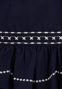 Tissu marine avec surpiqûres blanches brodées, présentant des rangées de motifs en pointillés, en croix et en points au-dessus d'une section froncée.