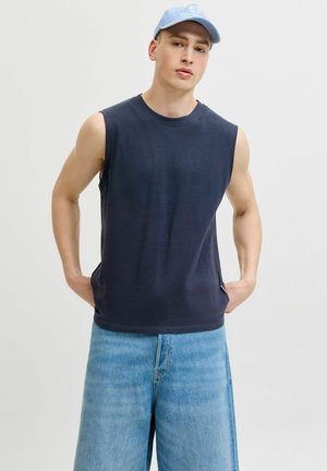 Jeune homme portant une casquette bleu clair, un débardeur bleu marine et un jean denim bleu clair à jambes larges, debout les mains dans les poches.