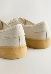 Baskets basses en toile beige avec des semelles épaisses en caoutchouc marron clair, vues de dos sur une surface blanche.