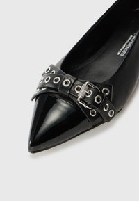Ballerines noires à bout pointu en cuir verni brillant, corps en cuir texturé, et un nœud avec œillets en métal argenté et une boucle métallique.