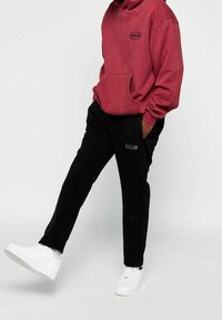 Bordowa bluza z kapturem z przednią kieszenią i logo, zestawiona z czarnymi spodniami z boczną kieszenią na zamek, uzupełniona białymi trampkami.